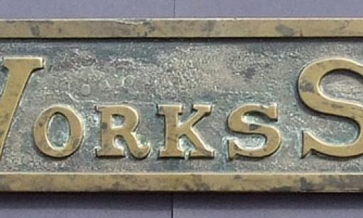 Letters and numbers engraved on a slightly tarnished bronze plaque with holes on each end.