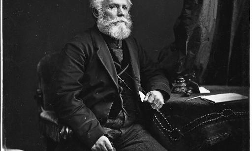 A well-dressed man with a thick white beard sits with his arm resting on a nearby table.