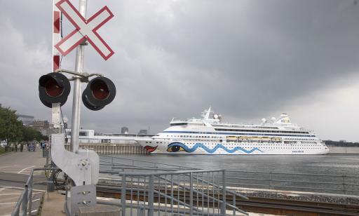 View from next to a railroad of people strolling by the river in the distance, near a cruise ship moored at a wharf.
