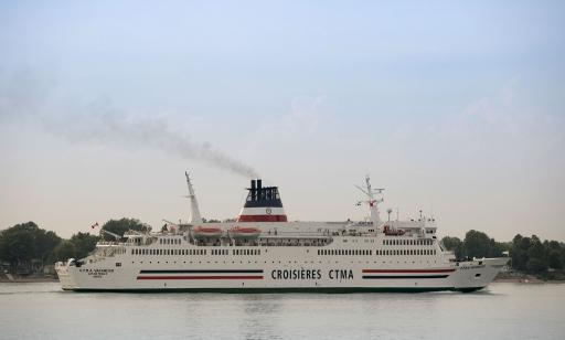 A large white ship, with one blue and one red stripe, sails near a shore lined with trees and houses.