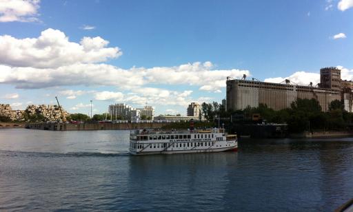 Un navire blanc se déplace sur l’eau en direction du canal Lachine et au loin on voit des tours d’habitations et des silos.