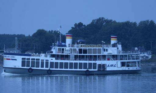 In the early evening, a white ship with yellow, red and blue accents sails near the shore.