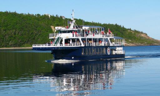 Un bateau bleu et blanc navigue devant une montagne. À l’avant de ce navire, le pont inférieur s’avance au-dessus de l’eau.