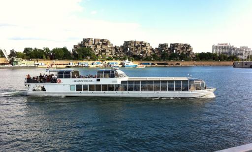 A boat with a glassed-in main deck sails on the river in front of a housing complex made of superimposed cubes.