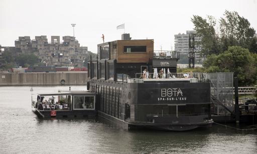 Un bateau noir avec une rallonge sur le côté est amarré près d’un parc. Au loin, on voit une habitation en blocs superposés.