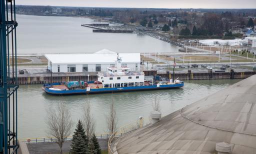 View from a tower of cars and trucks on a flat boat that is pulling up in front of a building on a wharf.