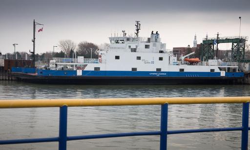 A flat blue and white boat, loaded with cars, is moored at a wharf. The steeples of a church can be seen in the distance.