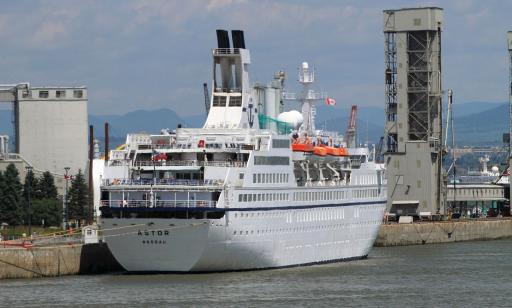 Stern view of a very large white ship moored at a wharf, showing its tall stacks and many decks.