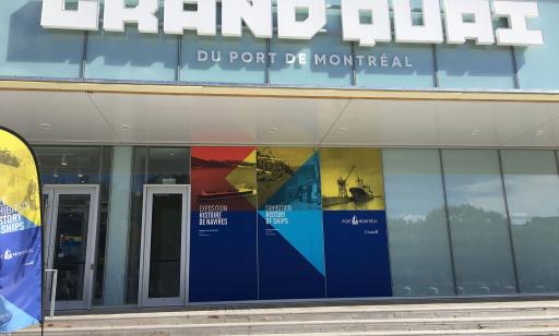 Sur le devant d’un édifice vitré, on voit l’inscription Grand Quai du Port de Montréal et une affiche bleue, jaune et rouge.