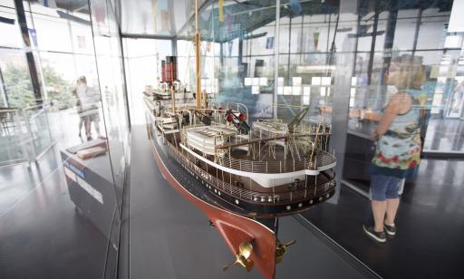 Close-up of a model ship in a glass-walled room, with visitors in the background.