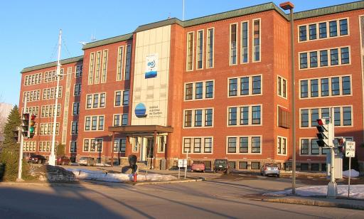 A red brick building with many windows. On its façade is a logo shaped like the letter Q filled in with blue bands.