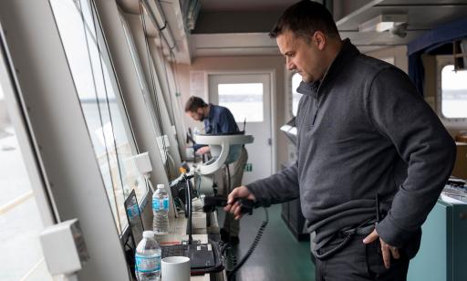 Two men, one near and the other farther away, consult their computers in front of the large windows of the wheelhouse.