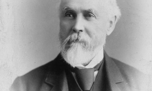 Black and white bust portrait of a bearded man with white hair, wearing a dark vest and jacket.