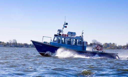 Three men in the cabin of a blue and white boat moving quickly on the water. Houses on the shore can be seen in the distance.
