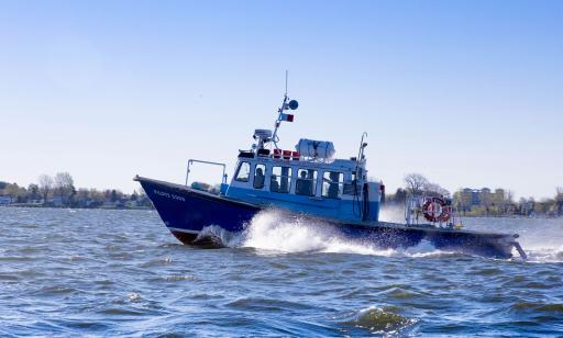 Three men in the cabin of a blue and white boat moving quickly on the water. Houses on the shore can be seen in the distance.