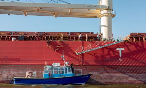 A metal ladder with rope railings is lowered alongside the hull of a cargo ship toward a man waiting on a boat.