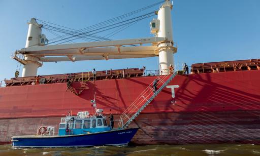 A man walks up a ladder lowered alongside the orange hull of a cargo ship. Another man stands at the bottom on a small boat.