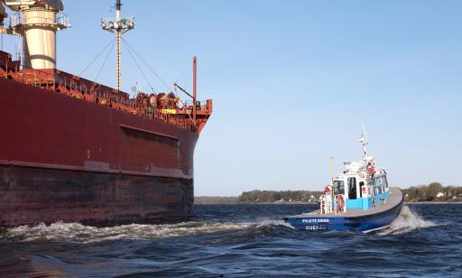 A blue and white boat speeds away from the prow of a huge cargo ship. Houses on the shore can be seen in the distance.