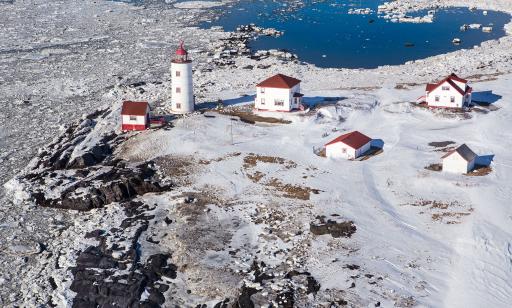 A lighthouse, two houses and three outbuildings with red roofs stand on rocky, snow-covered ground near the frozen river.
