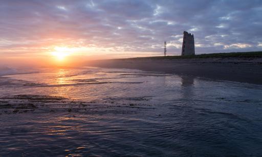 A metal skeleton tower and a lighthouse with a ruined top stand out against the purple sunset light.