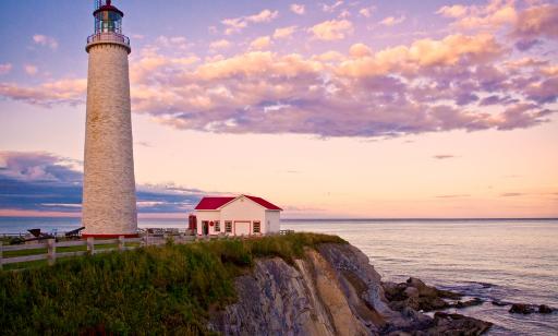 A lighthouse and a white house with a red roof stand atop a cliff. Below, water stretches out as far as the eye can see.