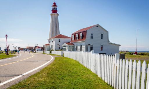 Several white buildings with red roofs stand along a road leading to a very tall white lighthouse topped with a red lantern.