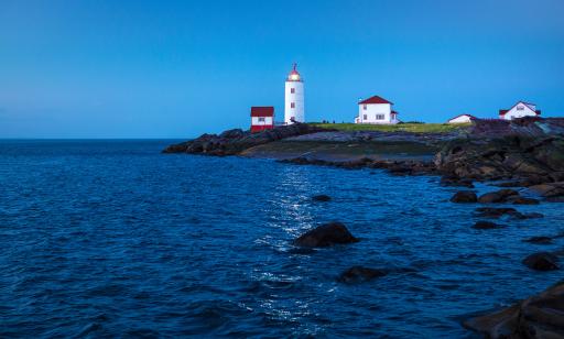 The water reflects the light shining from a white lighthouse with a red lantern room standing on a rocky shore.