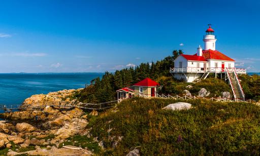 A white house with a red roof and a built-in lighthouse stands atop a rocky hill on an island, near a wood.