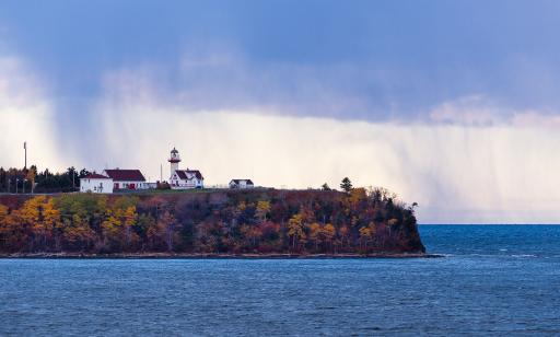 Above a bluff covered in trees showing their fall colours, a few buildings and a lighthouse stand on a cape under the rain.