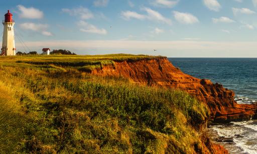 Des bâtiments blancs aux toits rouges et un phare blanc dont la lanterne est rouge sont en haut d’une falaise de grès rouge.