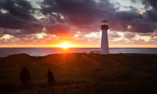 A man and a woman walk near a lighthouse under the golden light of the sun as it sets over the wide waters of the gulf.