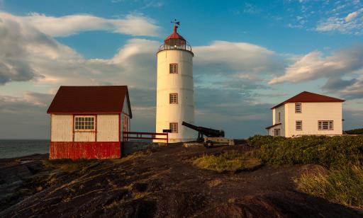 On a rocky outcrop where rose bushes grow stand a white lighthouse, two cannons, a small outbuilding and a house.