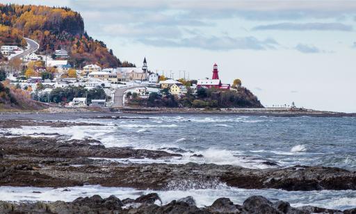 A lighthouse looks out over the gulf from atop a bluff. Light snow lies on the ground and the trees show their fall colours.