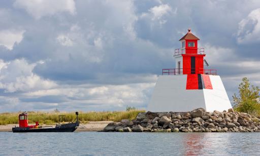 A white lighthouse on a mound of rocks. A wide red band with a narrow black band in the middle is painted on the front.