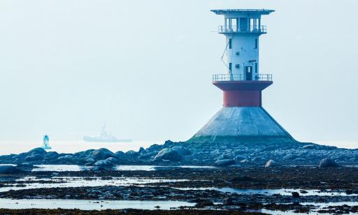 At low tide, the base of a lighthouse built on the rocky bottom can be seen. A tug sails through the fog in the distance.