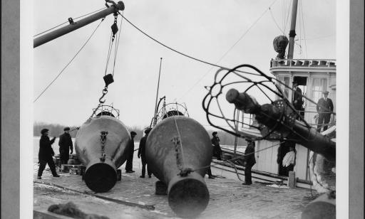 Men on a wharf use hoists to lift two large metal buoys.