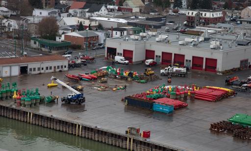 Sur un quai où sont empilées des bouées jaunes, rouges et vertes, on voit des camions et une grue qui soulève une bouée.