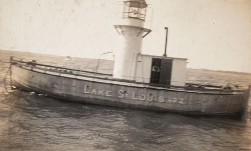Un bateau, ancré au milieu d’un cours d’eau, porte un phare octogonal au milieu et une cabine qui lui est accolée.