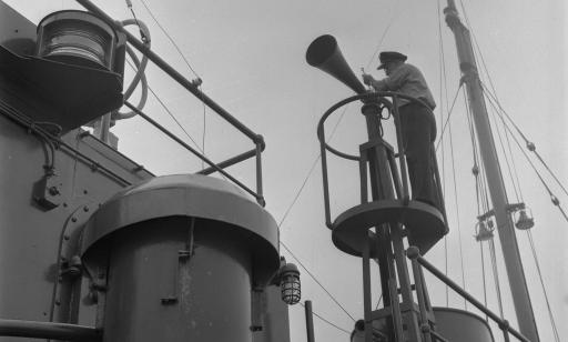 A man wearing a cap stands on top of a turret on a ship, next to a foghorn.