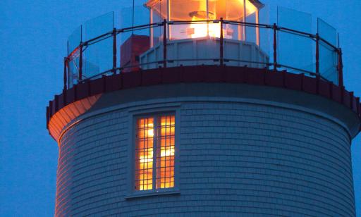 En soirée, on voit le haut d’un phare recouvert de bardeaux en bandes rouges et blanches et la lumière dorée de la lanterne.