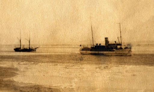 A steamship with a large smokestack uses a cable to tow a smaller boat with furled sails.