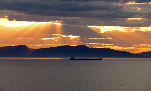 Les rayons orange du soleil traversent les nuages et illuminent la surface de l’eau où se découpe la silhouette d’un navire.