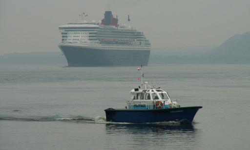 At dusk, a small blue and white boat sails away from a large ship with an enormous smokestack and many decks.