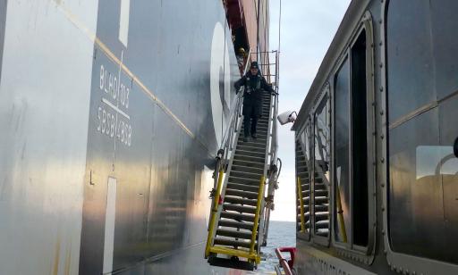 A man stands on a ladder being lowered from a ship to the deck of a smaller boat.