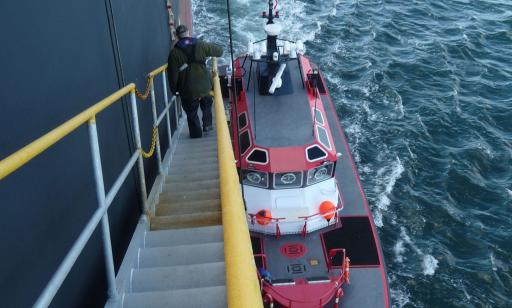 Back view of a man going down a metal ladder alongside the hull of a large ship to a small boat.