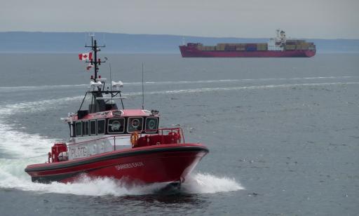 On voit de face un petit bateau rouge et blanc qui navigue en formant une vague et au loin un porte-conteneurs très chargé.