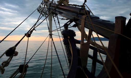 View of the prow of a large wooden sailing ship, with its bowsprit, figurehead and cables, and mountains in the distance.