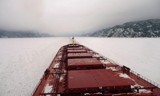 View from the wheelhouse of a cargo ship sailing on a frozen river, with snow-covered mountains on either side.