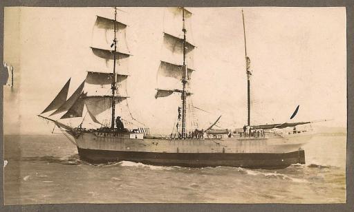 Black and white photo showing men climbing in the mainmast rigging of a three-masted sailing ship.