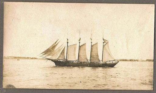 Black and white photo of a four-masted sailing ship on the river, with the shore in the distance. 